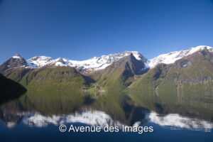 Majestic snow-capped mountains reflect perfectly on the calm, clear waters of Hjorundfjord. The lush greenery contrasts with the snow, under a vibrant blue sky.